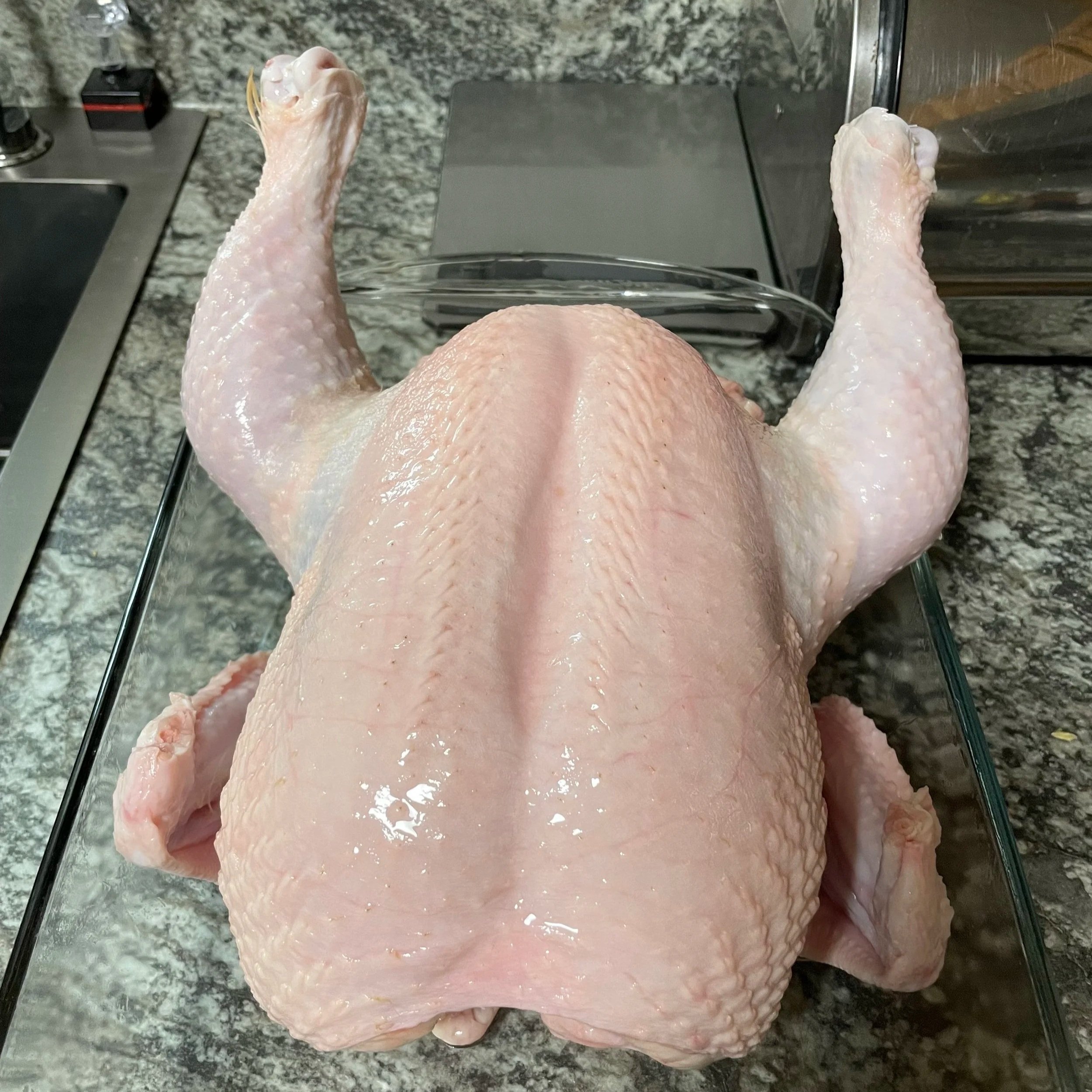 Raw whole chicken displayed on a glass dish, showing the back and wings, set on a granite countertop.