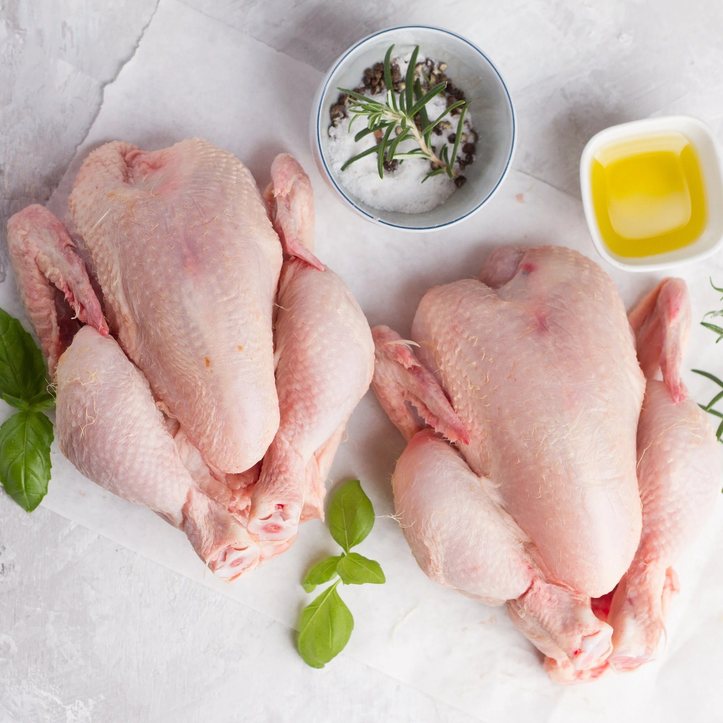Raw whole chickens placed on parchment paper, accompanied by a bowl of salt and herbs, and a small dish of olive oil, garnished with fresh basil leaves.