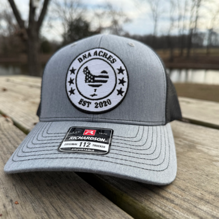 Gray trucker hat with a circular patch featuring an American flag and the text "DNA Acres EST 2020," resting on a wooden surface near a pond.