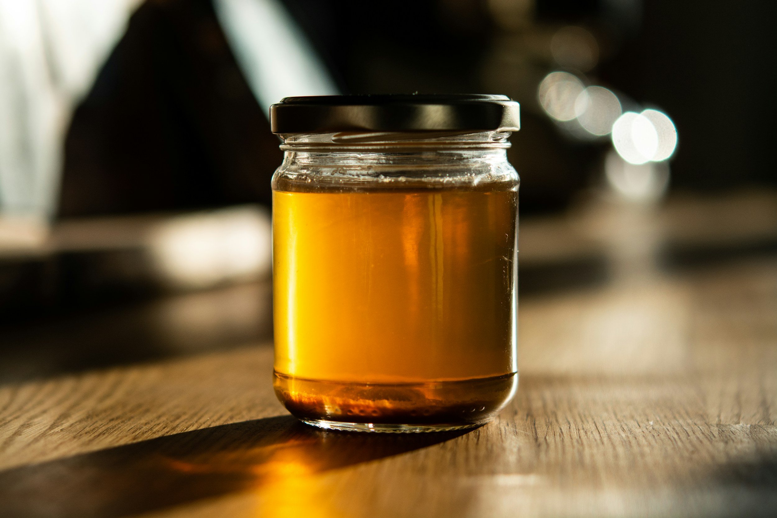 Jar of golden honey sitting on a wooden table, illuminated by soft natural light, showcasing its rich color and texture.