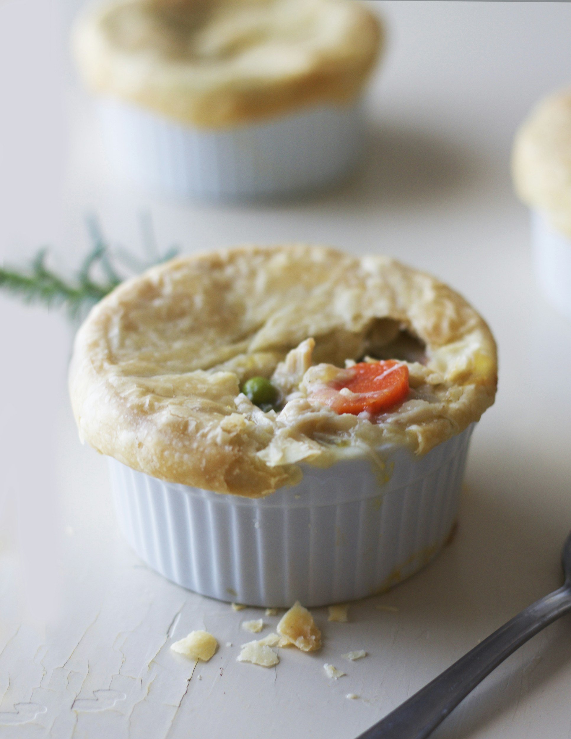 Individual chicken pot pie in a white ramekin, featuring a flaky golden crust and visible filling of chicken, peas, and carrots, with a spoon beside it on a textured surface.